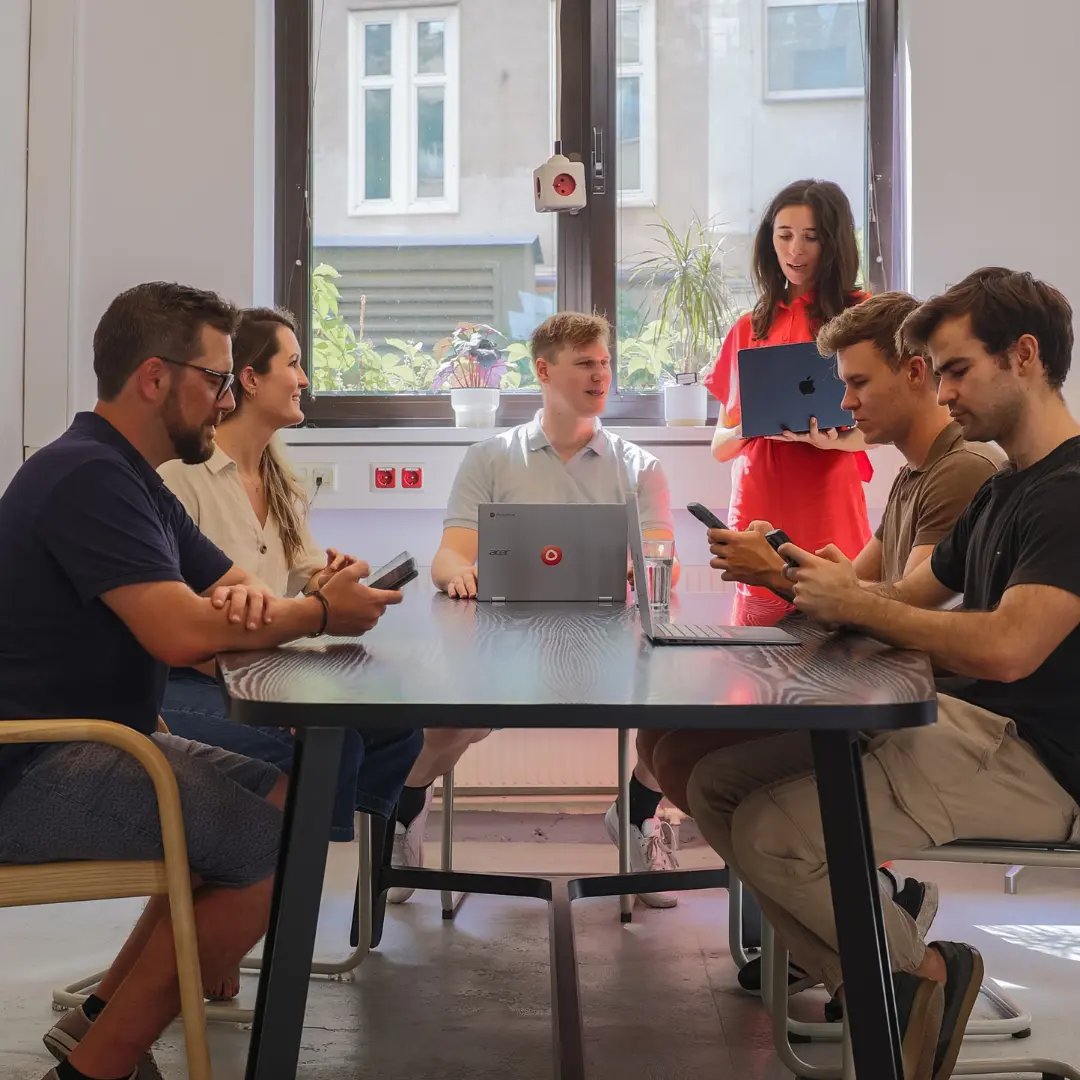 Sechs Mitglieder des Einfachmarketing-Teams sind in einem Büro um einen Tisch versammelt; einige sitzen mit ihren Telefonen, während einer mit einem Laptop in der Hand dasteht und interagiert.