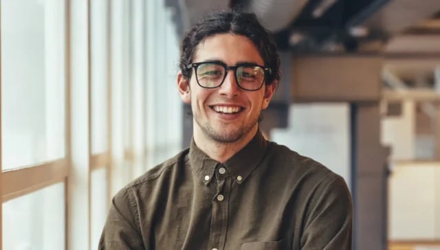 Ein Mann mit langen dunklen Haaren und Brille, der ein zugeknöpftes olivgrünes Hemd trägt, steht in einem Innenraum an großen Fenstern und lächelt in die Kamera - er strahlt das selbstbewusste Auftreten eines fraktionierten CMO aus.