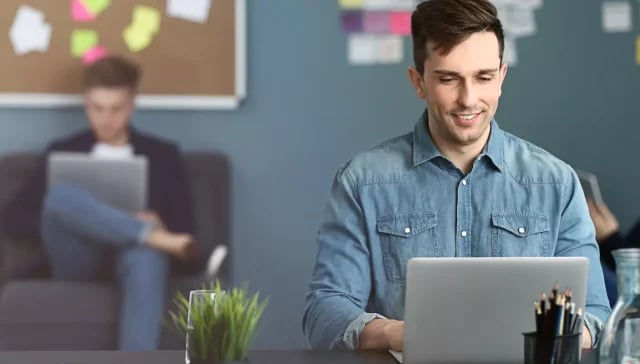 Ein Mann im Jeanshemd verwendet in einer geschäftigen Büroumgebung einen Laptop zur Marketingunterstützung, während im Hintergrund Kollegen arbeiten.