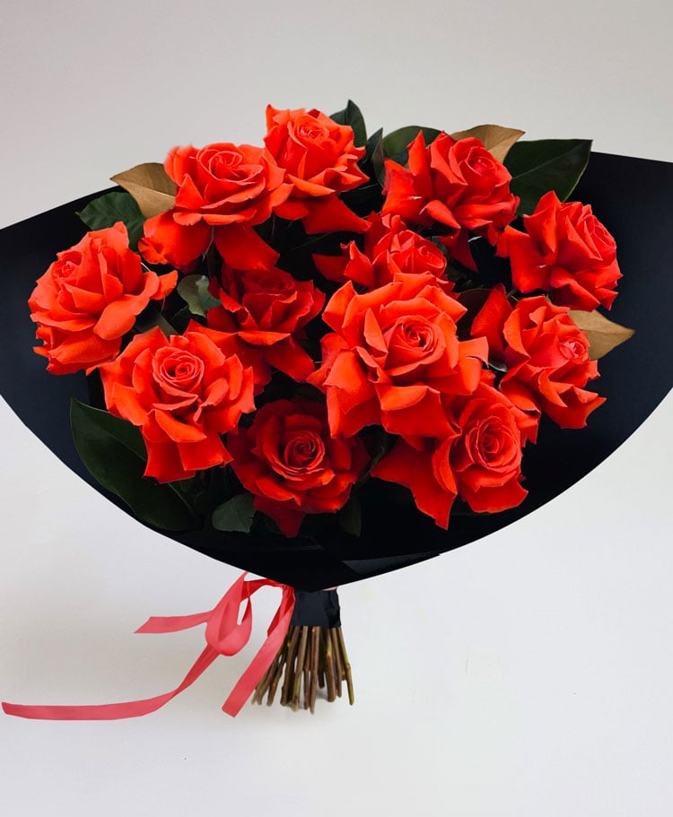 A bouquet of vibrant red roses and green leaves wrapped in black paper with a red ribbon, set against a plain white background, from the Dozen Roses - Wrapped collection.