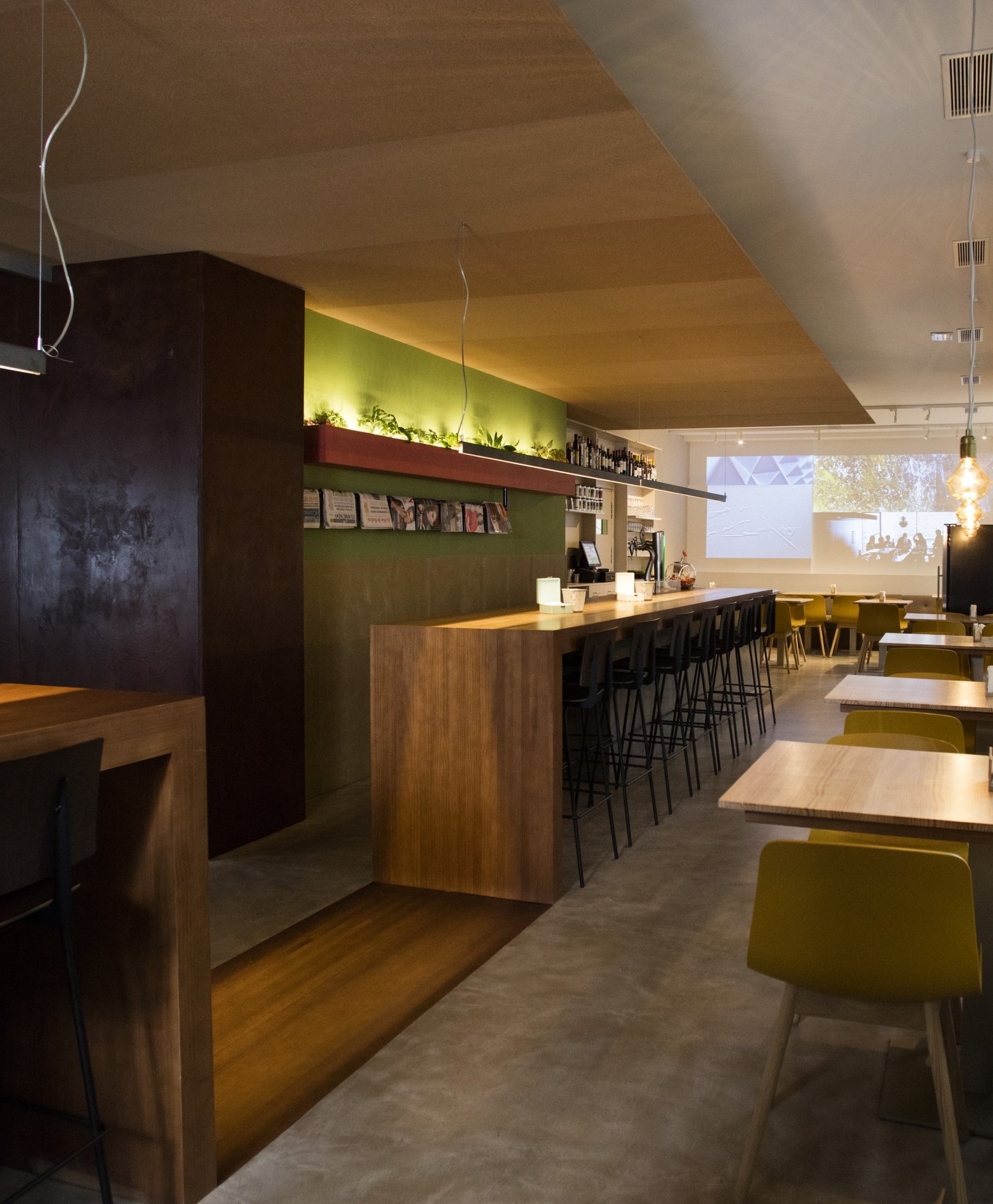 Interior de restaurante moderno con barra de madera, sillas amarillas y proyección en la pared – Un ambiente acogedor y contemporáneo para disfrutar de una comida o café en Santiago de Compostela.