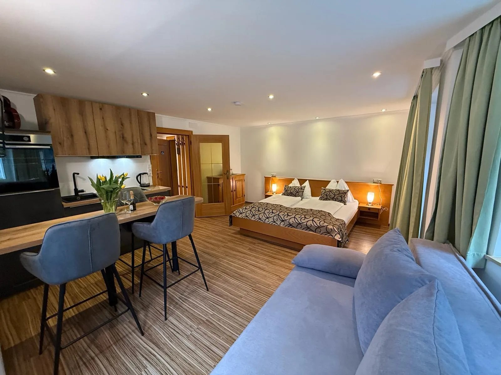 Studio-Apartment mit Doppelbett, gemütlichem Sofa, moderner Küchenzeile und Esstisch mit zwei Barhockern – großer Raum mit hellen Vorhängen und Blick ins Grüne.