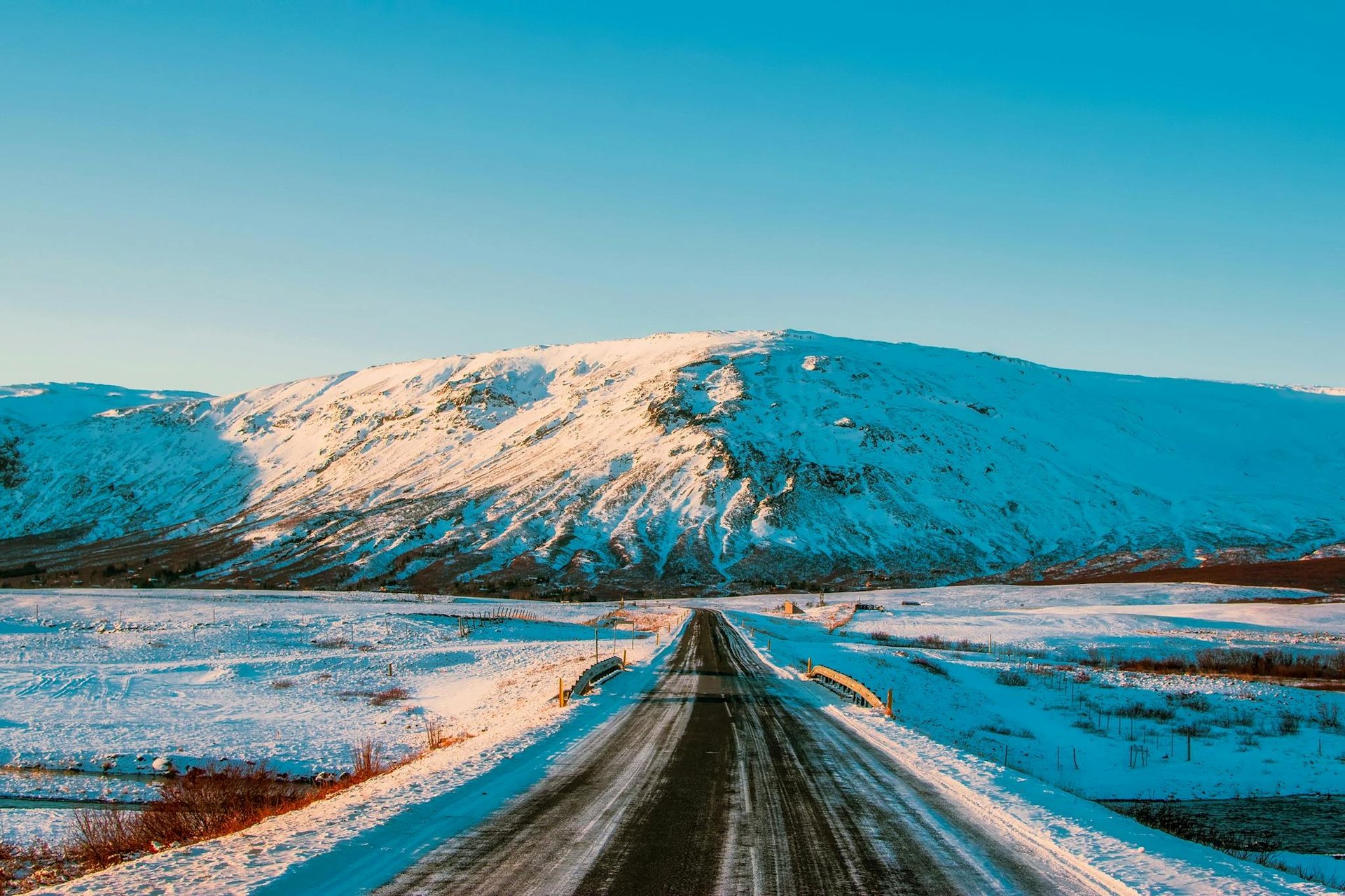 A picturesque winter road leading towards snow-capped mountains under a bright blue sky.