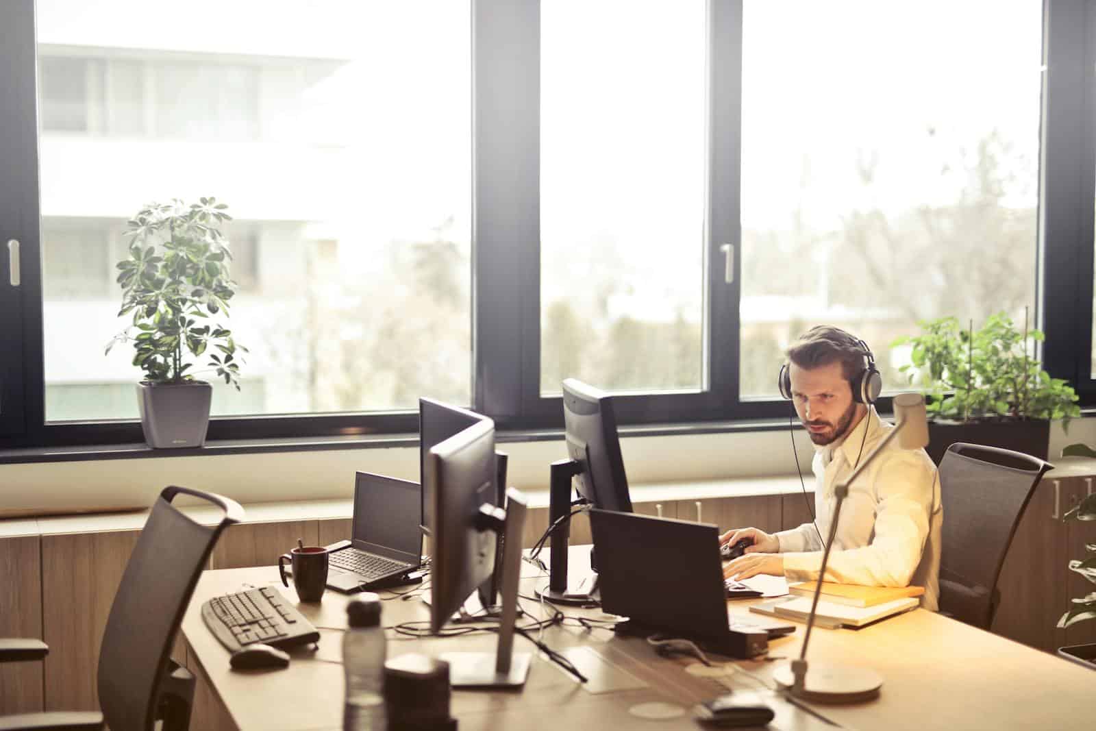 A businessman sits at a desk using multiple computers and a headset in a well-lit modern office. Hosting support for website design agencies