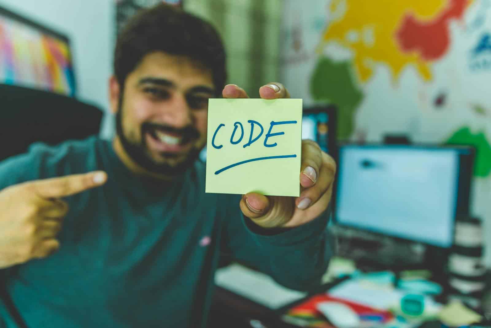 Programming languages, A smiling programmer pointing to a sticky note with 'code' written on it in an office setting.