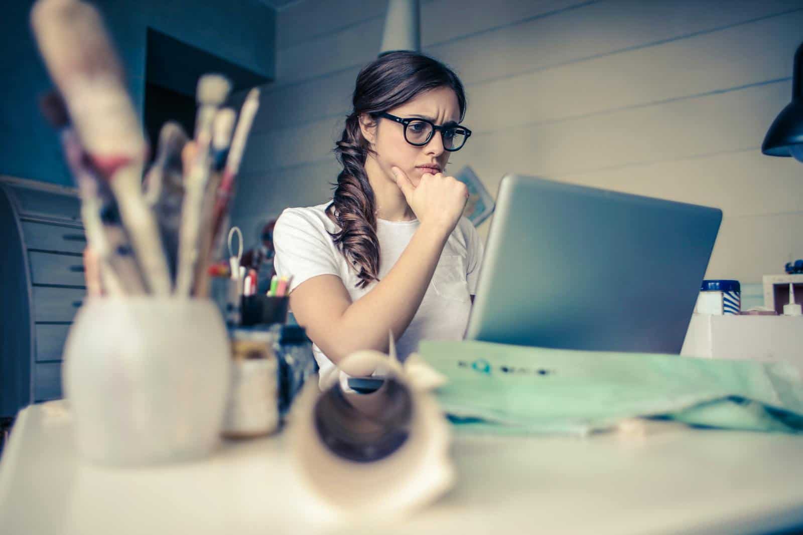 Optimizing LinkedIn Company Pages, Young woman with glasses deeply focused on a laptop surrounded by art supplies in a home office.
