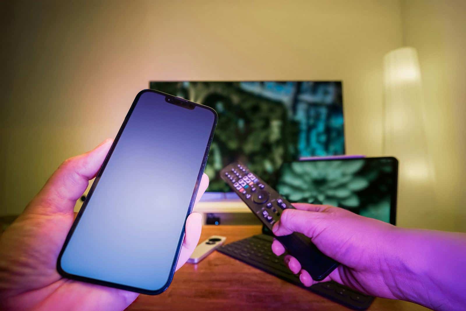 Brand Persona, communication Chanels, Hands holding a smartphone and remote control in a high-tech home setup with TV and other electronic devices.