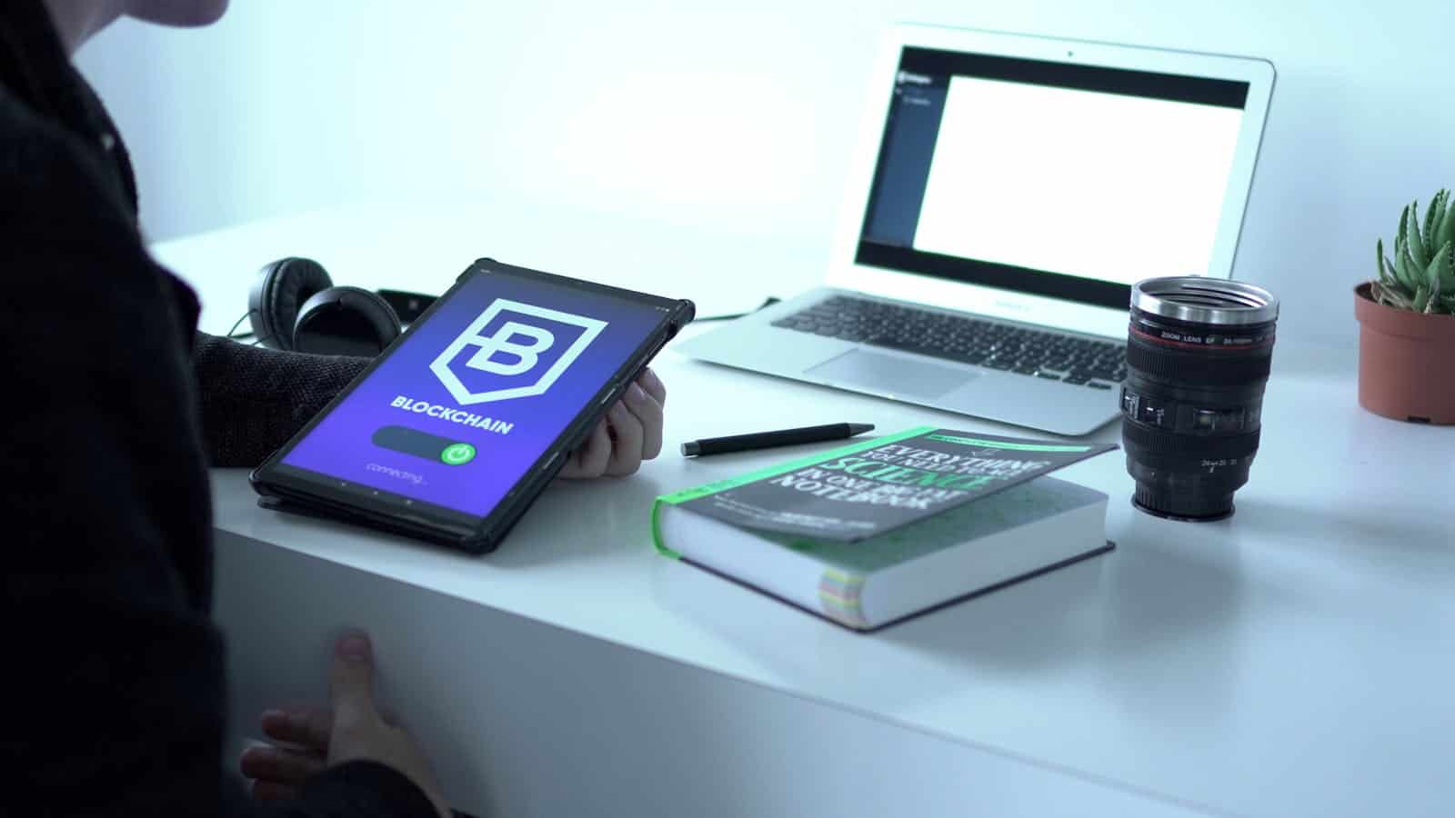 Website security. A desk with tablet displaying blockchain, laptop, book, and camera lens, symbolizing tech and innovation.