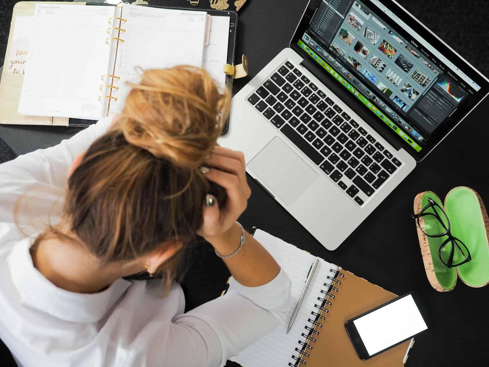 Overhead view of a stressed woman working at a desk with a laptop, phone, and notebooks. Lifetime subscribtion, lifetime deal, subscription fatigue, small business, productivity tools