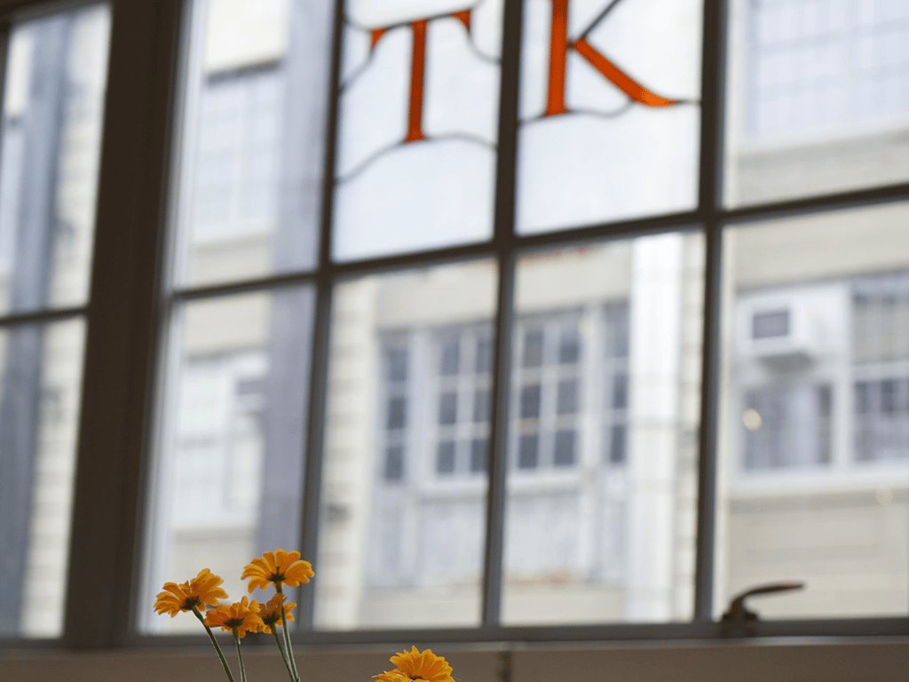 Small yellow flowers in a vase sit on a windowsill, exuding an optimistic charm. A large window behind them reveals a blurred view of urban buildings, while the letters "T" and "K" form part of a creative stained glass design at the top of the window.