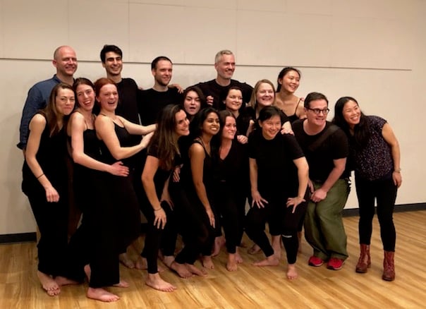 A group of sixteen people, wearing casual black attire, are gathered indoors on a wooden floor. They are smiling and posing together for a group photo. Some are standing while others kneel in front, embodying the relaxed and friendly atmosphere of their creative process at work.