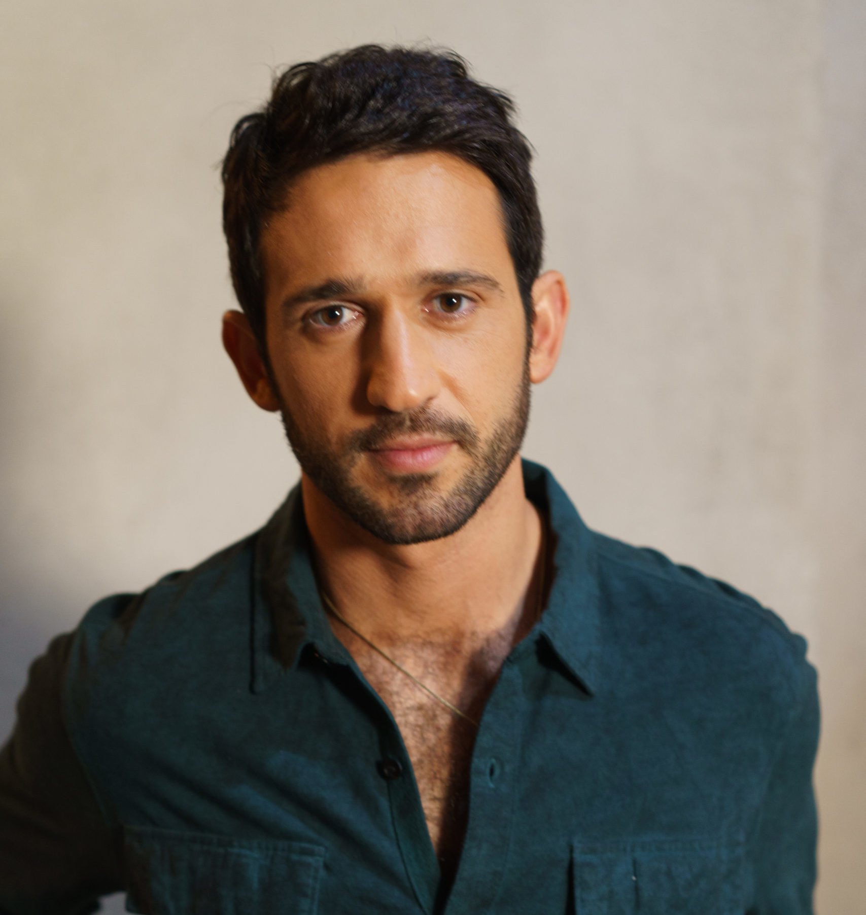 A person with short, dark hair and a beard is wearing a dark green shirt, reminiscent of a character from "Sex/Life 2" on Netflix. The plain, softly lit background frames their gaze as they look directly at the camera with a neutral expression.