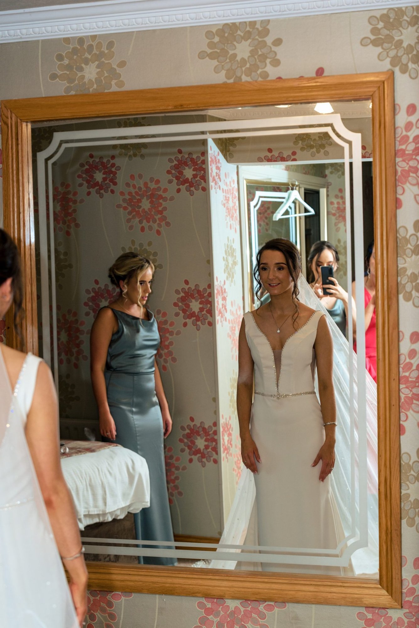 The Best Wedding venues - The Royal Court Hotel Portrush 3 Family Photographer A bride in a white dress viewing her reflection in a mirror at the Royal Court Hotel, accompanied by two women in formal attire.