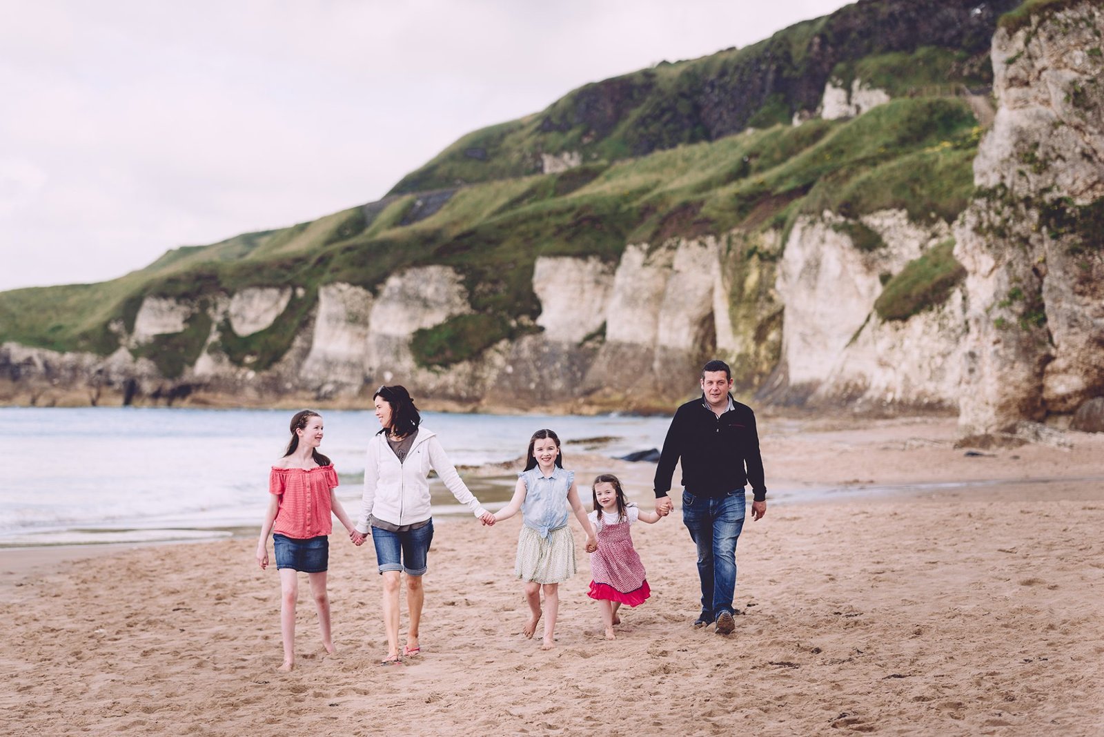 HOW NOT TO HAVE A NERVOUS BREAKDOWN AT YOUR FAMILY PORTRAIT SESSION! 3 Northern Ireland family photographers capturing a family walking on a beach with cliffs in the background.