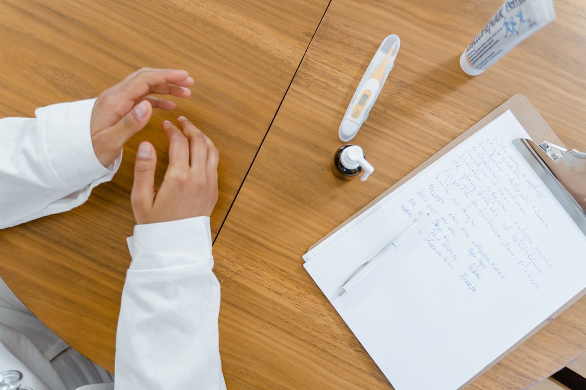 Person in a white coat sits at a wooden table with a clipboard of handwritten notes, a pen, a nasal spray, and a thermometer.