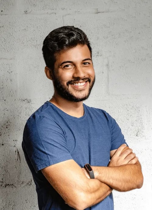 A man with short dark hair and a beard, wearing a blue t-shirt and a fitness tracker, stands with arms crossed and smiles in front of a white textured wall.