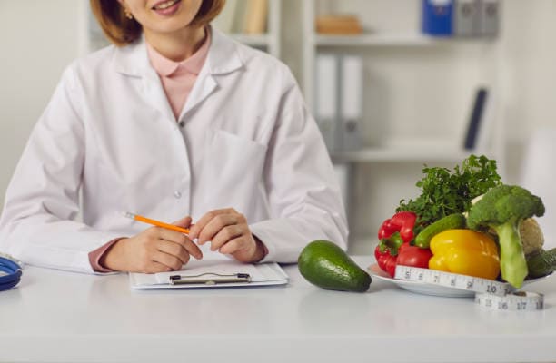 A person in a white lab coat sits at a desk with a clipboard, pen, avocado, assorted vegetables, and a measuring tape in front of them.