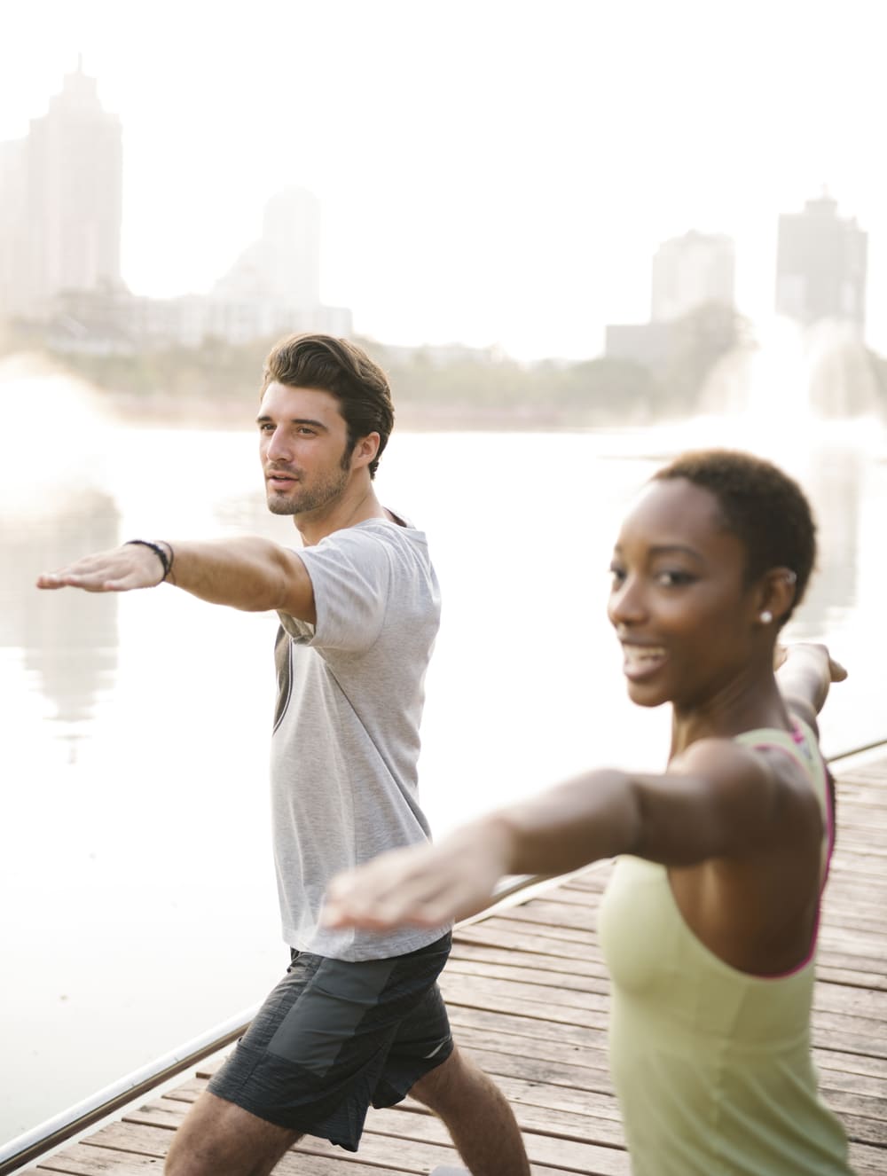 Two people stand on a wooden dock by the water, stretching their arms forward in a yoga pose with a cityscape and fountains in the background.