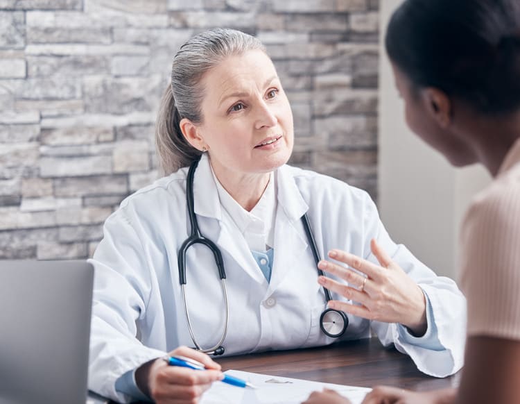 A doctor with a stethoscope talks to a patient across a desk, holding a pen and gesturing while discussing something.