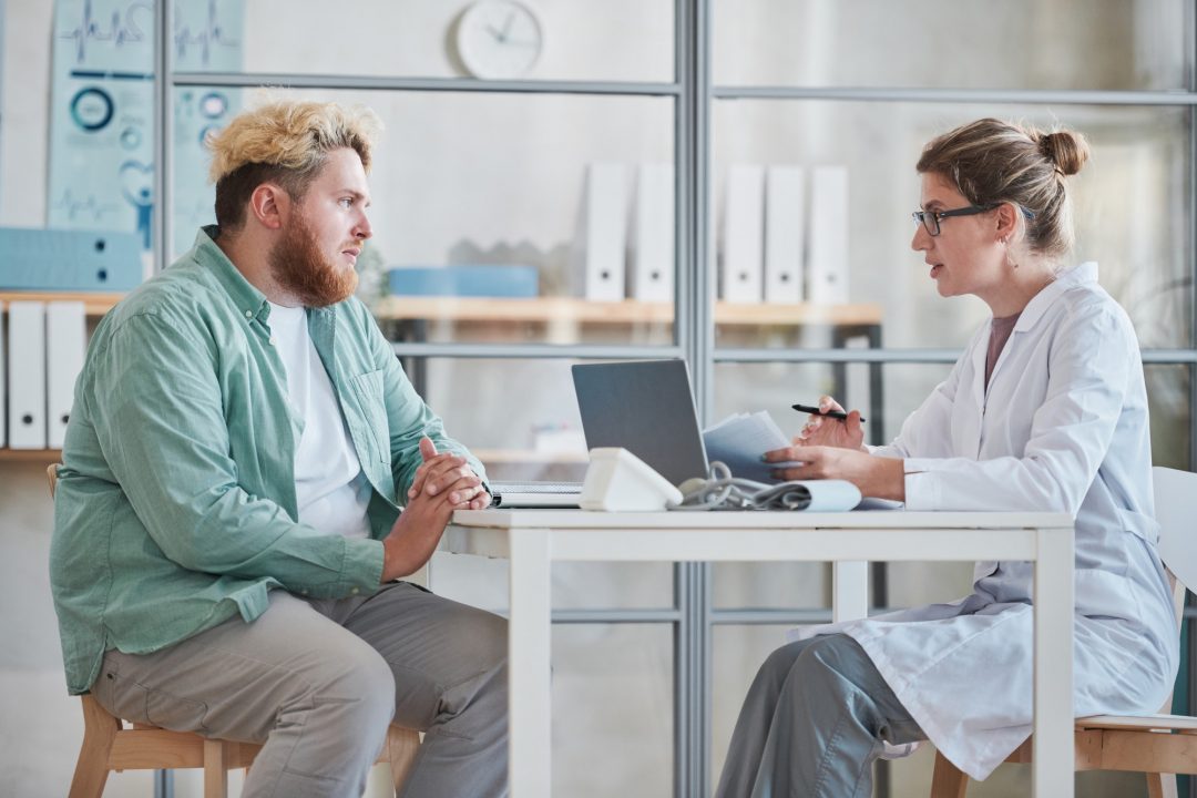 A doctor in a white coat consults with a patient sitting across from her at a table with a laptop and medical equipment in a bright office.