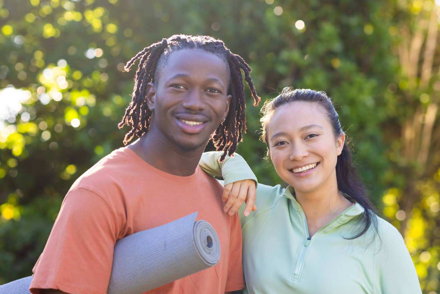 Two people standing outdoors; one holds a rolled yoga mat while both smile at the camera. Lush greenery is visible in the background.