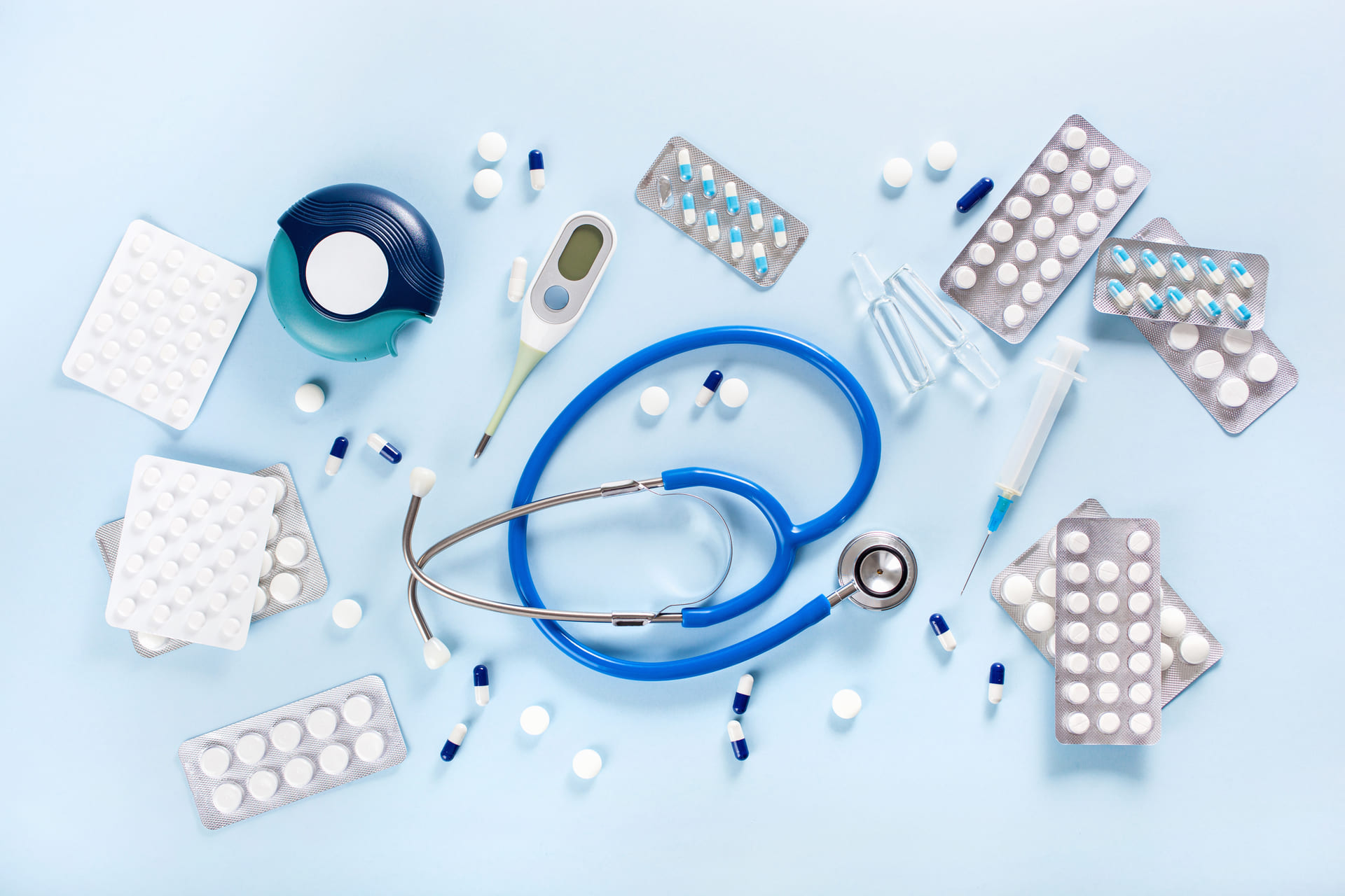 A stethoscope, thermometer, pill packs, loose capsules, syringes, and a tape dispenser arranged on a light blue surface.