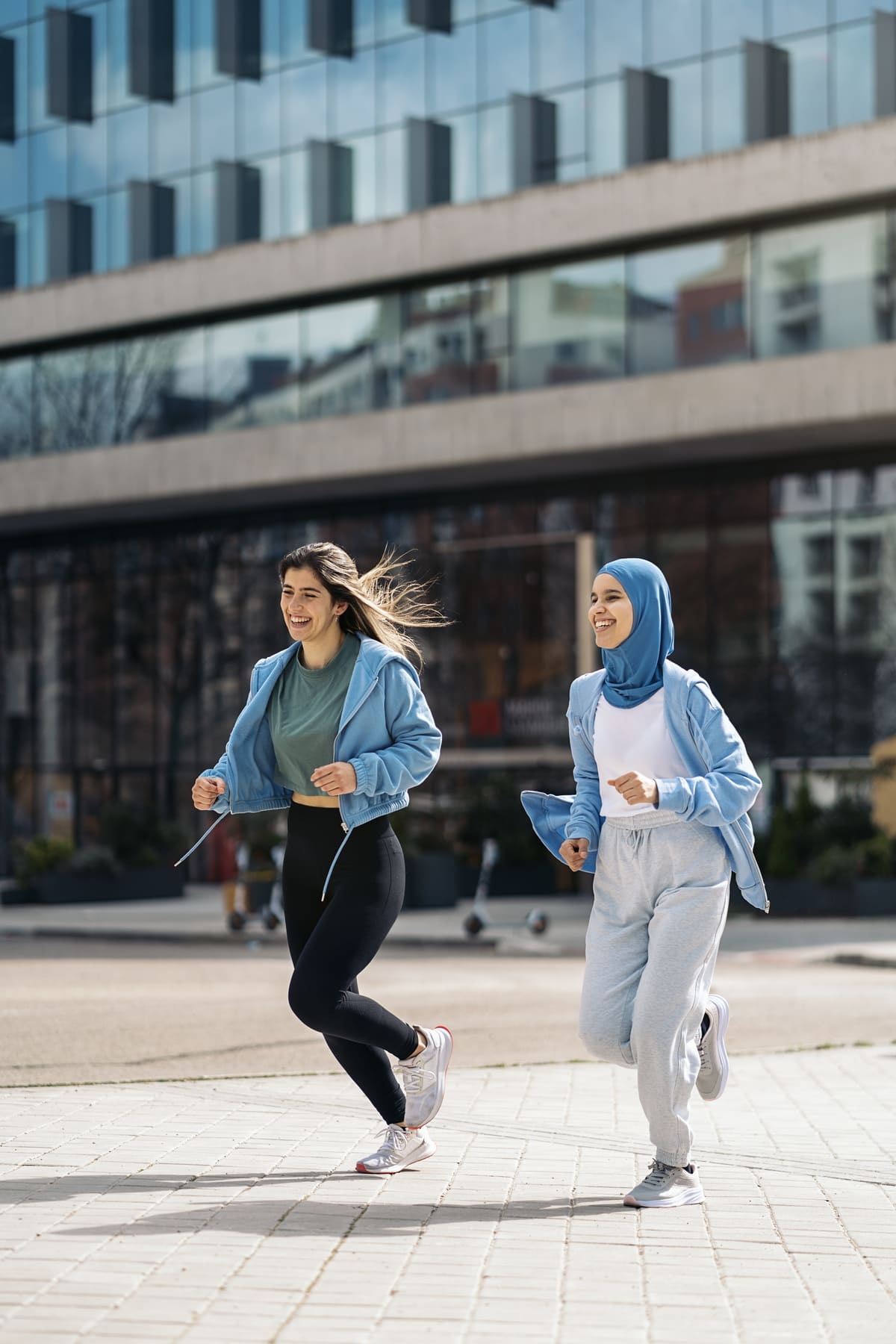 Two young women jogging outdoors in an urban area in front of a modern glass building, both wearing casual activewear.