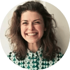 A woman with curly brown hair smiles broadly, wearing a green and white patterned shirt, standing against a plain light-colored background.
