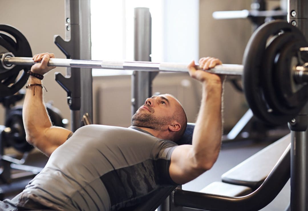 A man lies on a weight bench performing a barbell bench press in a gym, gripping the bar with both hands and preparing to lift.