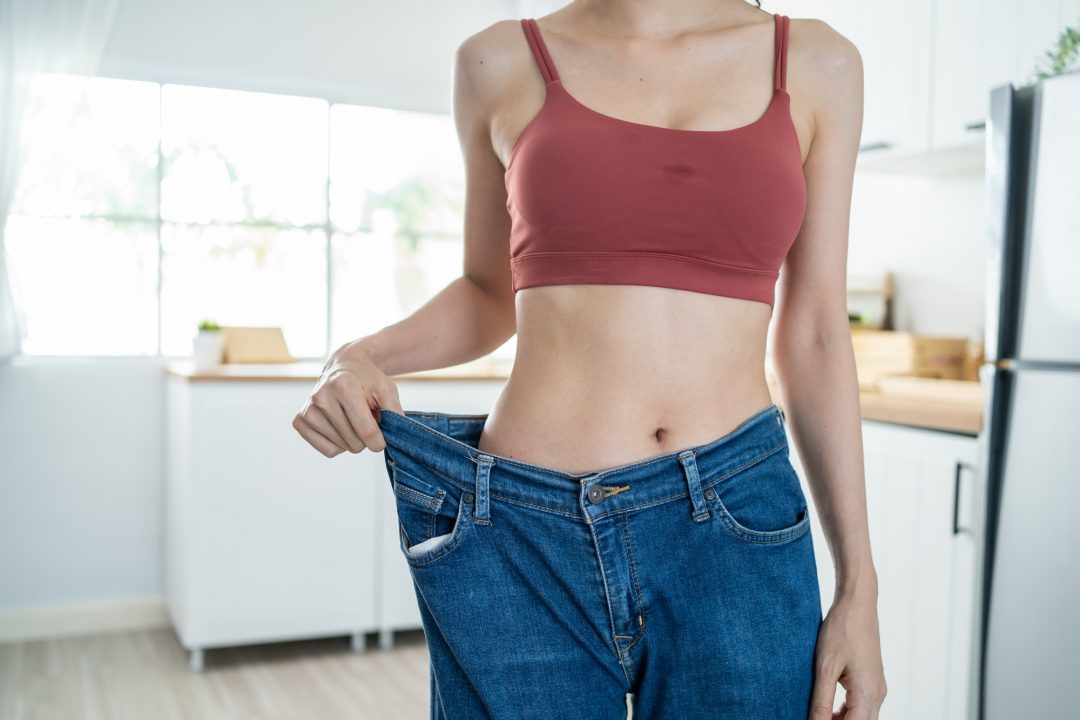 A person wearing a red sports bra stands in a kitchen holding out the waistband of loose blue jeans, suggesting significant weight loss.