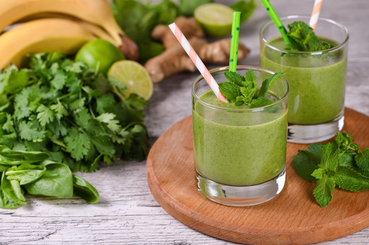Two glasses of green smoothie with straws and mint garnish on a wooden board, surrounded by bananas, greens, lime, and ginger.