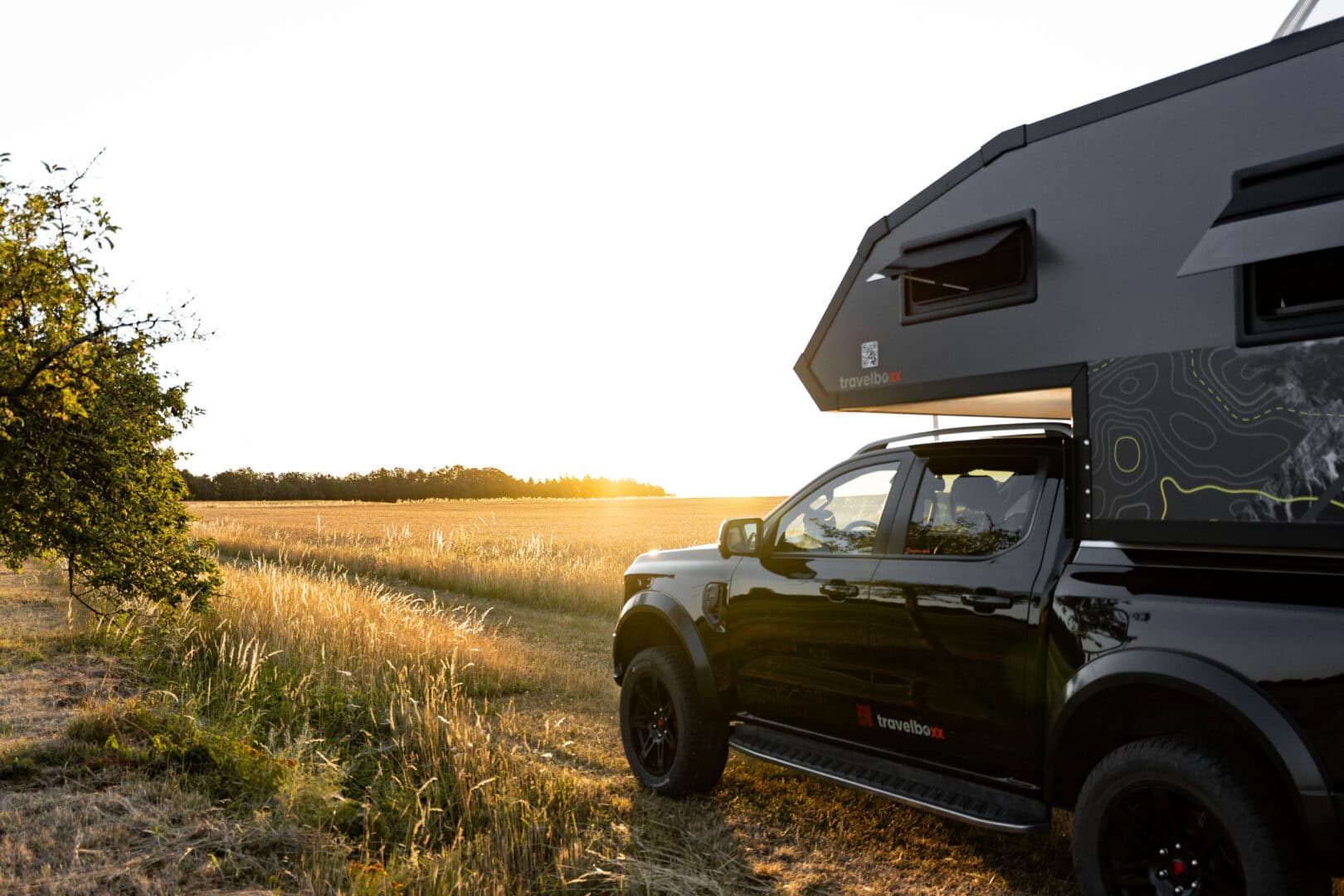 Ein schwarzer Pickup mit einem aufgebauten Camper steht bei Sonnenuntergang auf einer grünen Wiese. Das goldene Licht taucht die Landschaft und entfernte Bäume in eine stimmungsvolle Atmosphäre. Dieser Offroad-Camper kombiniert die Vielseitigkeit eines Pickup Campers mit individuellem, robustem Design - ideal für alle, die nach einem maßgeschneiderten Custom Camper für Abenteuer abseits befestigter Straßen suchen. Travelboxx - Tiny House Wohnkabinen