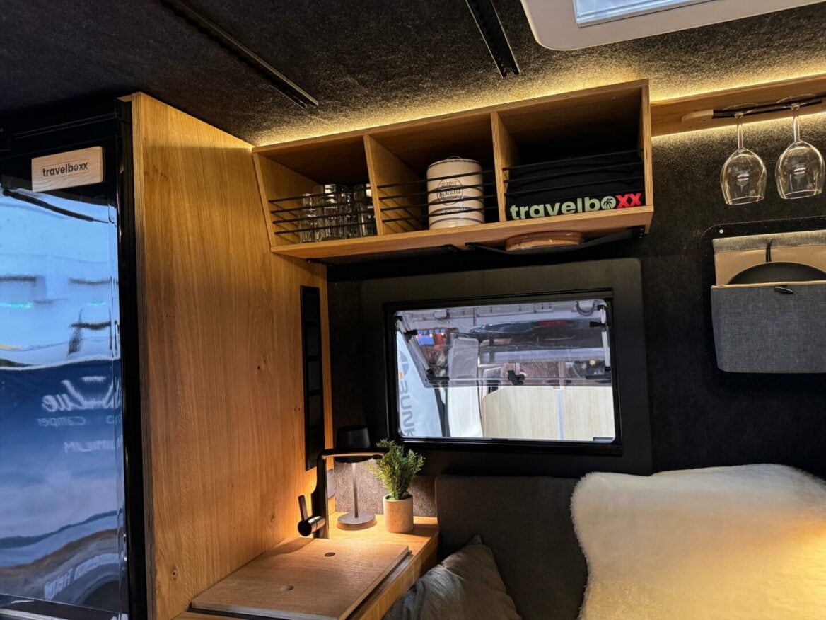 Gemütliches Pickup Camper-Interieur mit einem kleinen Schreibtisch aus Holz, Lampe, Pflanzen, Ablagen mit Geschirr und einer „Allround-Talent“ Travelboxx. Über dem eleganten Custom-Camper-Innenraum hängen Weingläser, während durch das Fenster grenzenlose Freiheit und Offroad-Abenteuer in der Natur sichtbar werden – ideal für individuelle Camper-Träume und anspruchsvolle Reisende auf der Suche nach dem perfekten Offroad-Camper.. Travelboxx - Pickup Wohnkabinen