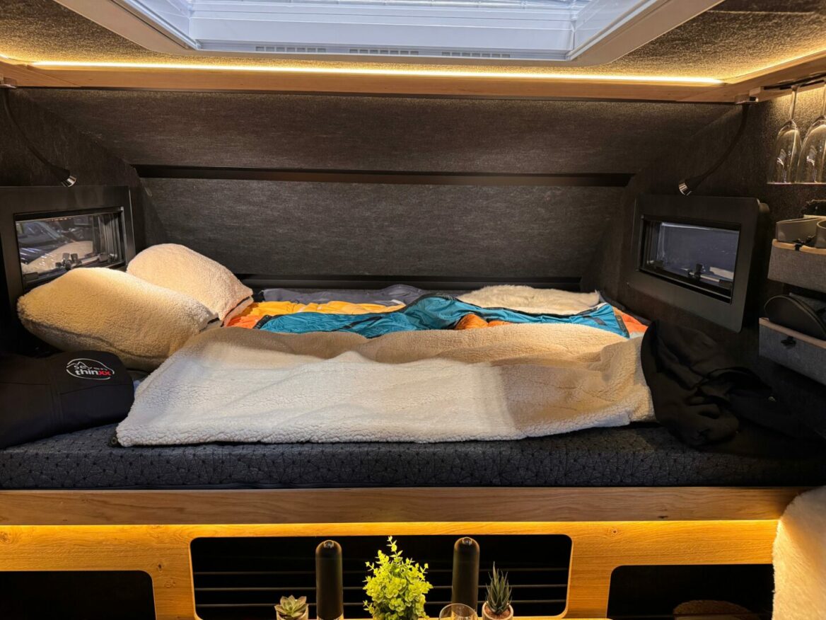 Gemütliches Bett-Setup im Camper – ausgestattet mit bunter Decke, bequemen Kissen, stimmungsvoller Beleuchtung und kleinen Fenstern auf beiden Seiten – deine PickupBoxx für grenzenlose Freiheit. Ein Regal darunter ist liebevoll mit kleinen Topfpflanzen dekoriert und verleiht deinem Offroad Camper oder Custom Pickup Camper eine wohnliche Atmosphäre, egal wohin dich dein Abenteuer führt. Travelboxx - Pickup Wohnkabinen