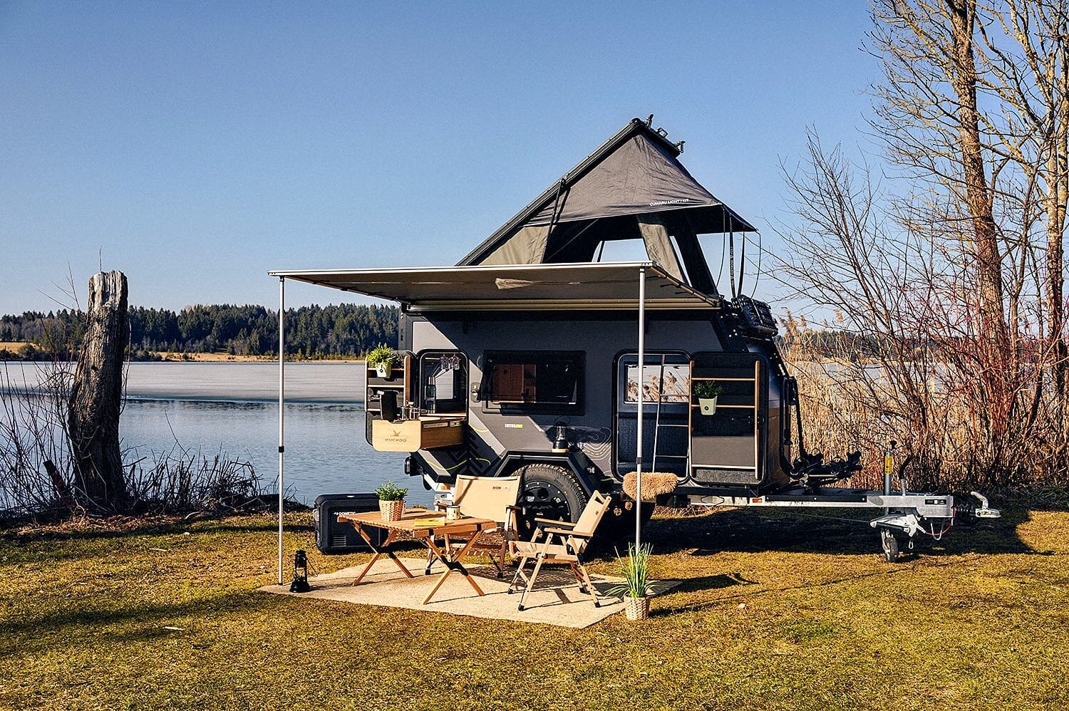 Ein moderner Travelboxx Offroad-Camper mit Aufstelldach steht idyllisch an einem See. Eine Markise verbringt Schatten über einem gemütlichen Outdoor-Essbereich mit zwei Stühlen, einem Tisch und Topfpflanzen. Umgeben von Bäumen und ruhigem Wasser wird dieser Custom Camper zur idealen Tiny House Oase für Individualisten, die einen Pickup Camper suchen und unabhängig in der Natur unterwegs sein möchten. Travelboxx - Pickup Wohnkabinen