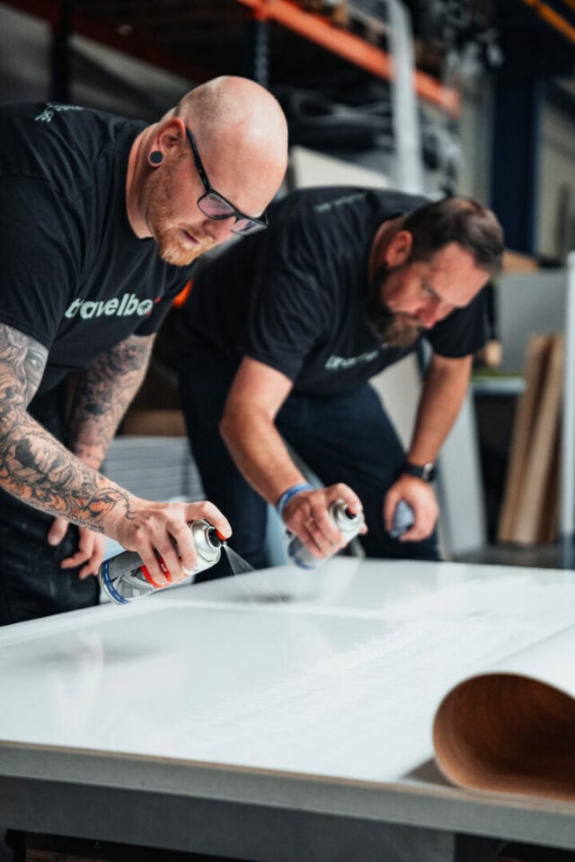 Zwei Männer in schwarzen Hemden und mit Brillen sprühen gemeinsam eine große, weiße Fläche in einer Werkstatt. Sie konzentrieren sich auf ihre Arbeit und halten jeweils eine Sprühdose in der Hand, während sie sich über einen Arbeitstisch lehnen. In dieser industriellen Umgebung werden maßgeschneiderte Pickup Camper oder Offroad Camper gefertigt - perfekt für alle, die auf der Suche nach einem individuellen Custom Camper sind. Travelboxx - Tiny House Wohnkabinen