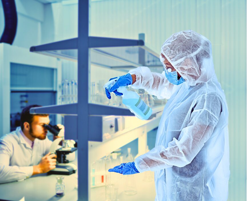 A Reinigungskraft in protective clothing sprays disinfectant in a Reinraum laboratory while another scientist observes samples through a microscope in the background.