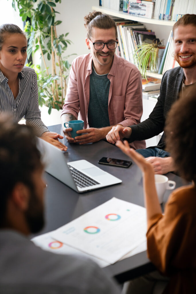 En grupp på fem personer sitter runt ett bord på ett kontor och diskuterar. En bärbar dator och dokument med färgglada diagram ligger på bordet, och i bakgrunden finns bokhyllor och växter. winwinweb