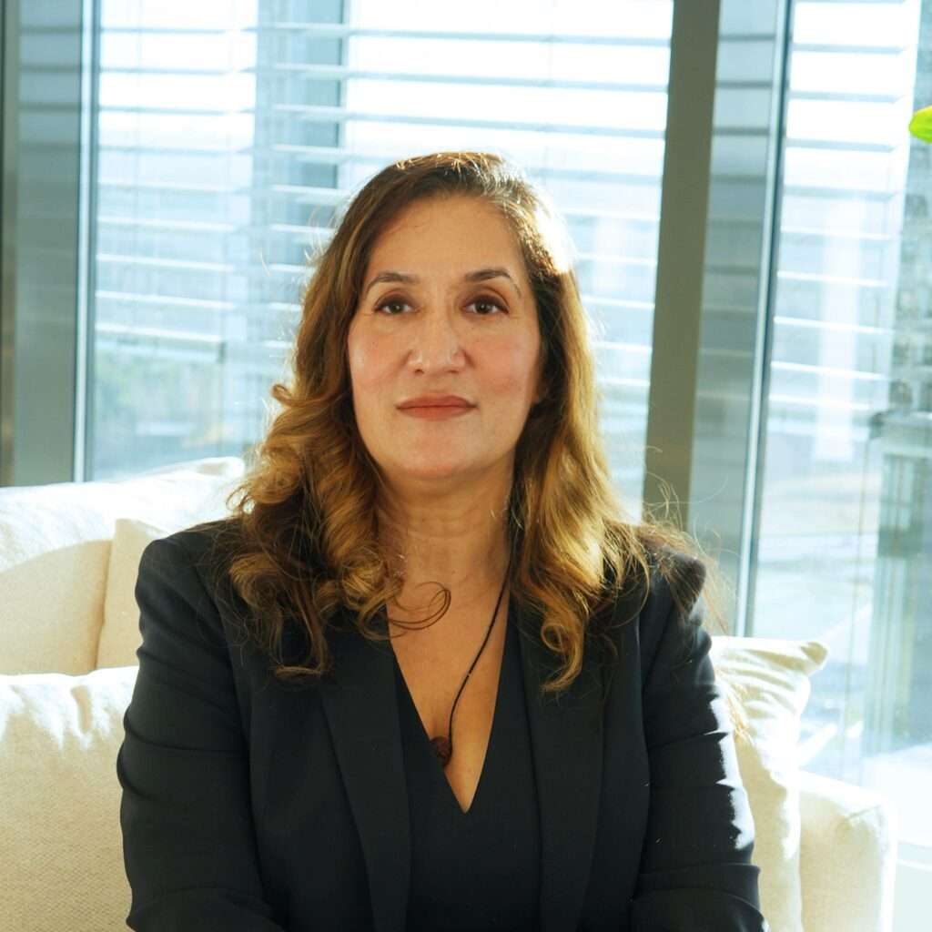 Jyotsna sitter i en ljus soffa i ett modernt rum med stora fönster och persienner. Solljuset lyser mjukt upp hennes ansikte. winwinweb