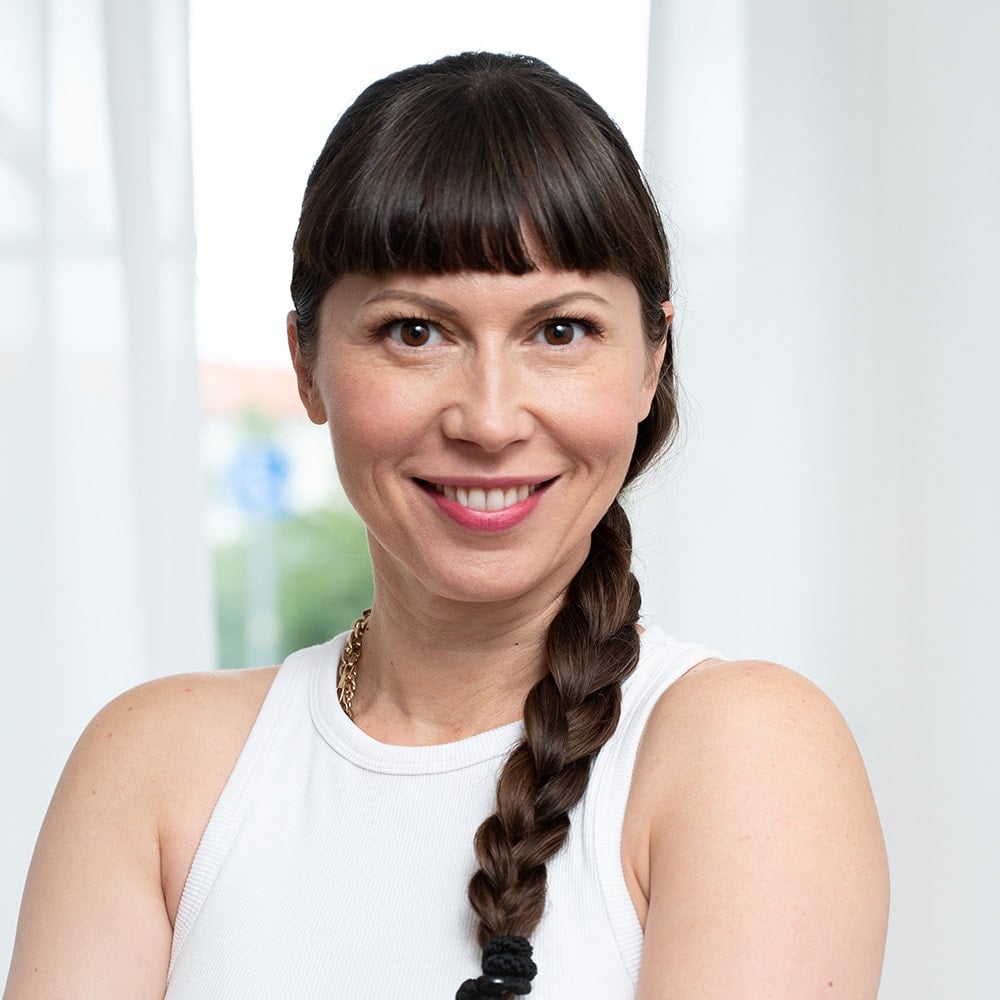 Maya, klädd i en vit ärmlös topp, ler mot kameran. Vita gardiner och en mjuk utomhusscen i bakgrunden.