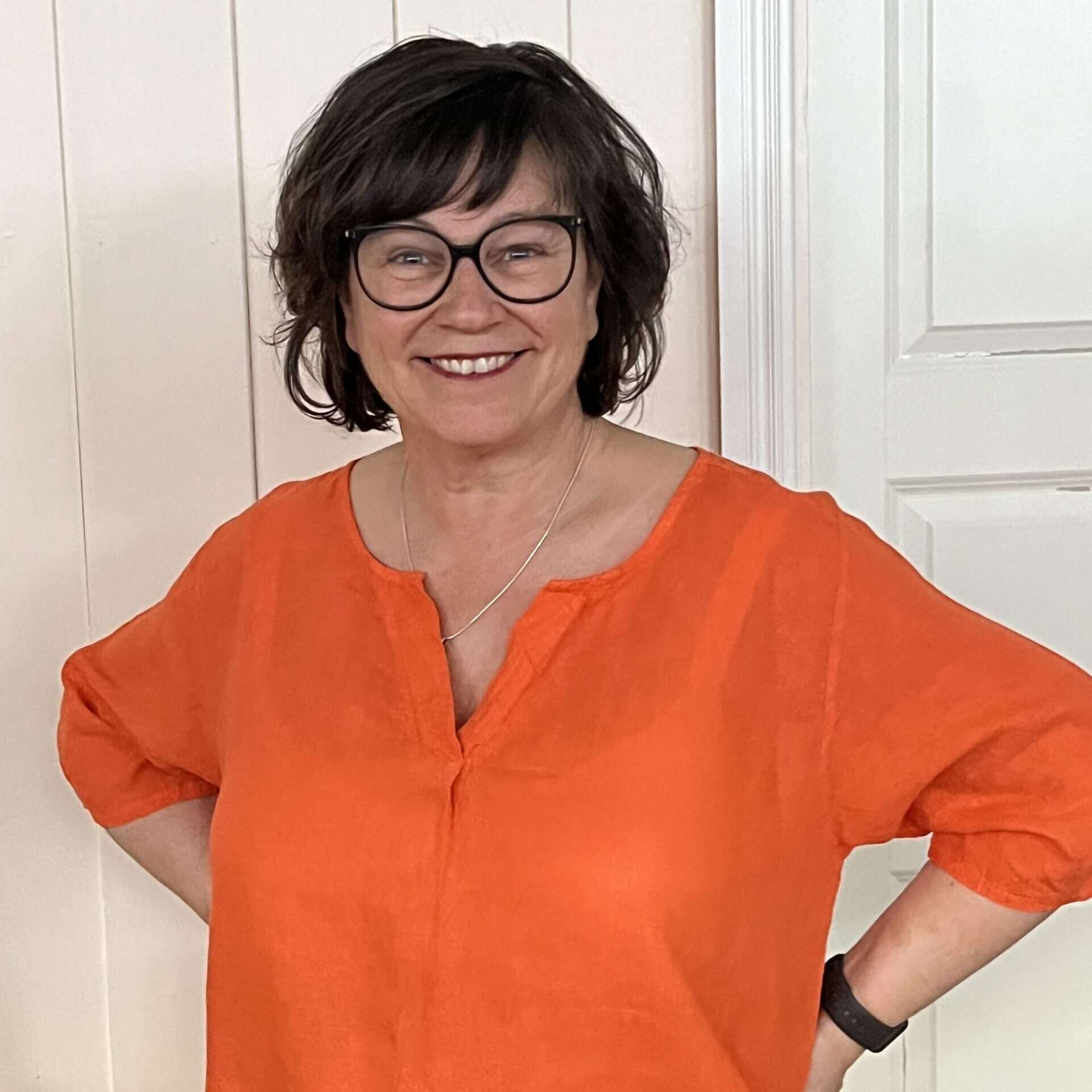En kvinna med kort mörkt hår och glasögon ler, stående med händerna på höfterna. Hon har på sig en orange topp och ett halsband och står framför en gräddfärgad vägg och en vit dörr. winwinweb